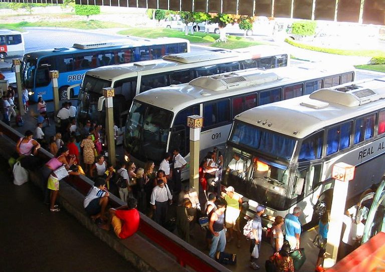 A foto mostra a imagem de ônibus interestaduais e seus passageiros em uma rodoviária