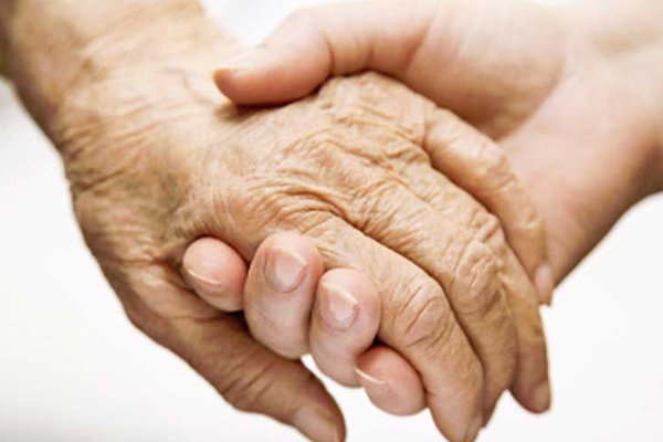 A imagem mostra uma mão de uma pessoa idosa sendo segurada pela mão de uma pessoa mais jovem