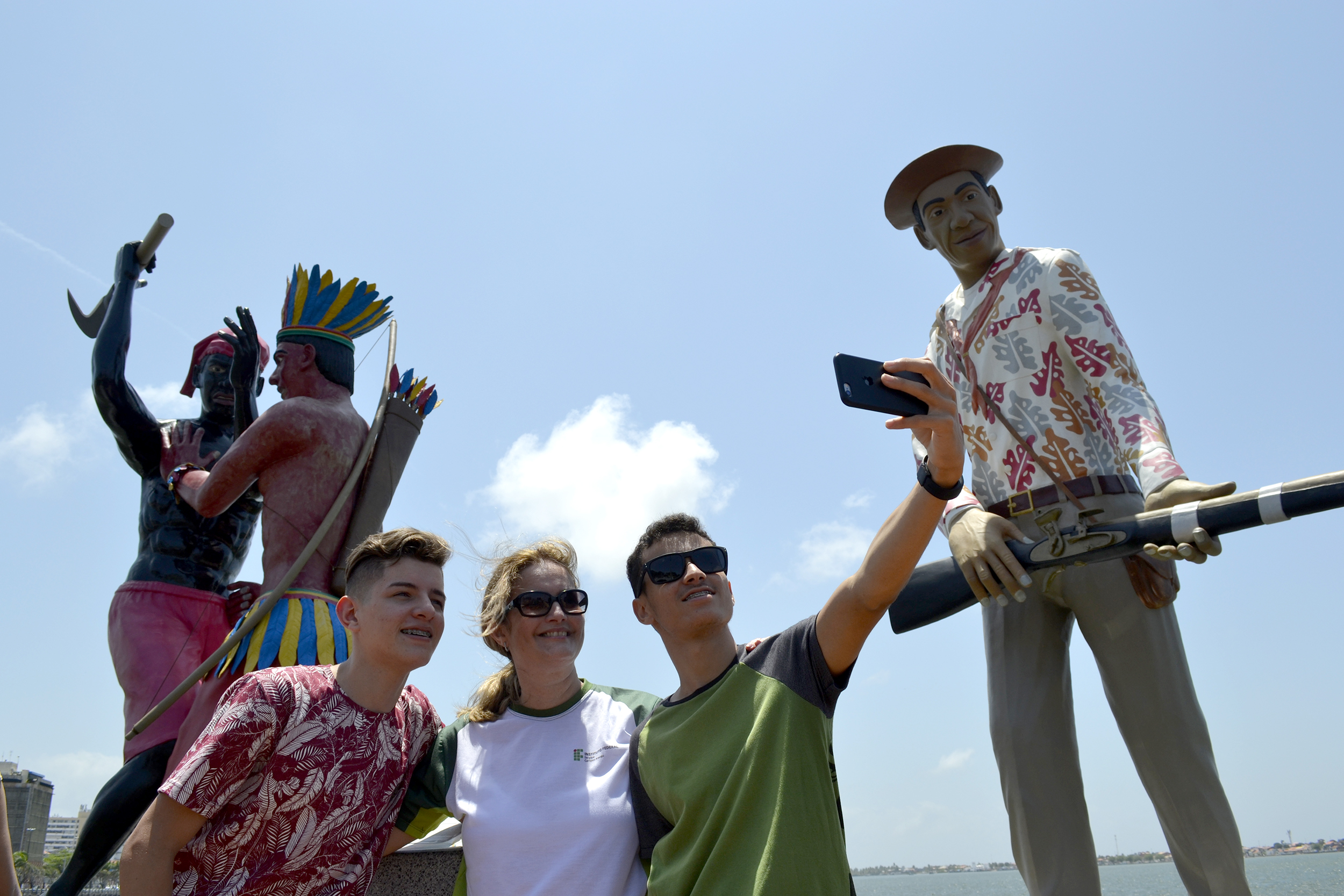 #PraCegoVer - Uma mulher faz selfie entre dois jovens. Ao fundo, esculturas do Largo da Gente Sergipana