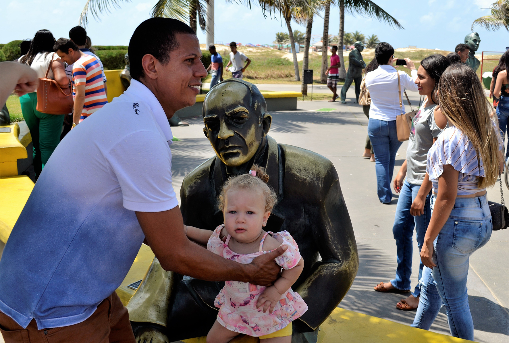 #PraCegoVer - Na Orla de Atalaia, homem coloca filha no colo de uma estátua para foto