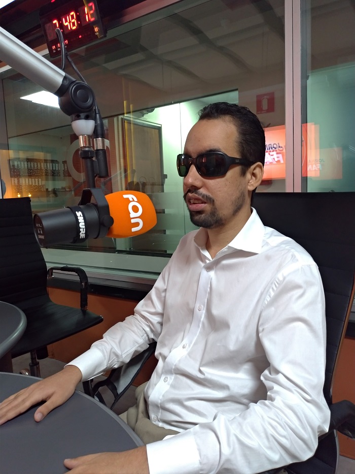 #PraTodoMundoVer Lucas Aribé sentado no estúdio da rádio, usando camisa branca. À frente dele está um microfone laranja com o nome FAN na cor branca