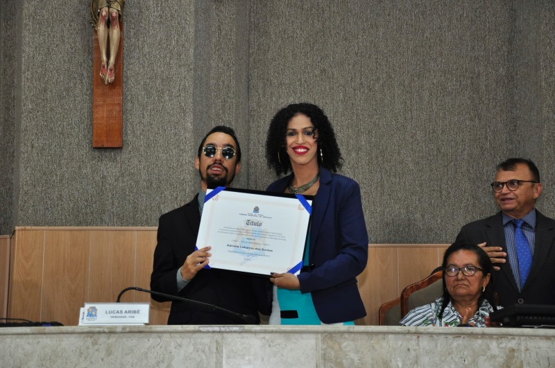 #PraTodoMundoVer Fotografia de Lucas Aribé ao lado de Adriana Lohanna na Mesa Diretora da Câmara segurando o diploma de cidadania