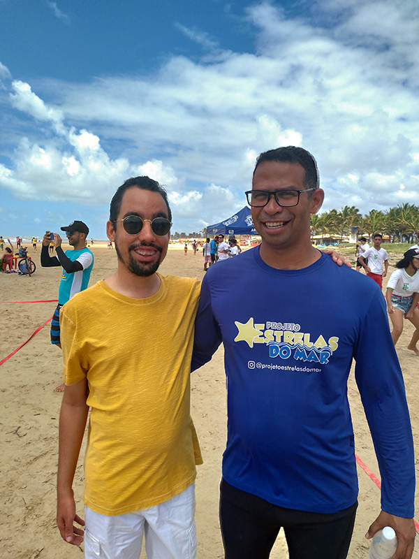 #PraTodoMundoVer Lucas Aribé, usando camisa amarela, em pé ao lado de Byron, usando camisa azul com marca do projeto Estrelas do Mar. Eles estão na areia da praia