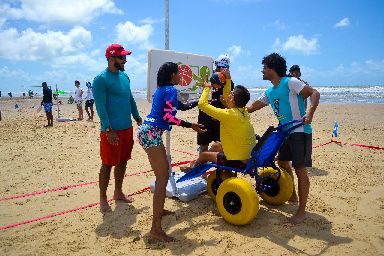 #PraTodoMundoVer Na areia da praia, cinco pessoas jogram basquete. Quatro homens e uma mulher, sendo que um dos homens usa cadeira de rodas adaptada