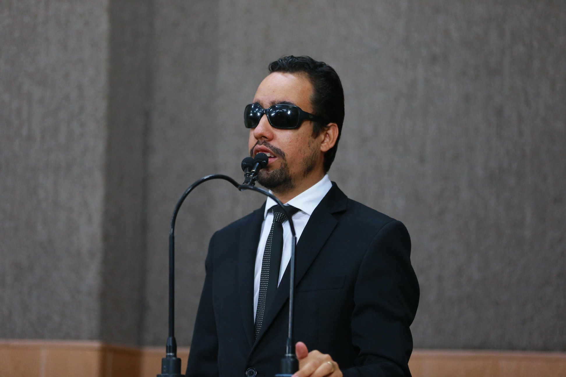 #PraTodoMundoVer Lucas Aribé em pé na tribuna falando ao microfone. Ele usa terno e gravata pretos e camisa branca. (Foto: César de Oliveira)