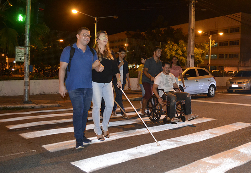 #PraTodoMundoVer Sete pessoas sobre uma faixa de pedestres. Uma mulher está com os olhos vendados, bengala, acompanahda de um guia, e um homem usa cadeira de rodas