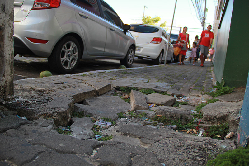 #PraTodoMundoVer Em primeiro plano, buraco na calçada com tijolos soltos. Ao fundo, carros estacionados ao lado da calçada, uma mulher, uma criança e um homem caminhando na calçada.