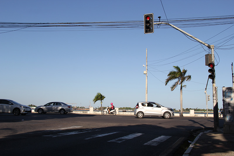 #PraTodoMundoVer Foto de uma avenida, onde há uma faixa de pedestres pintada e, acima, um semáforo com luz vermelha