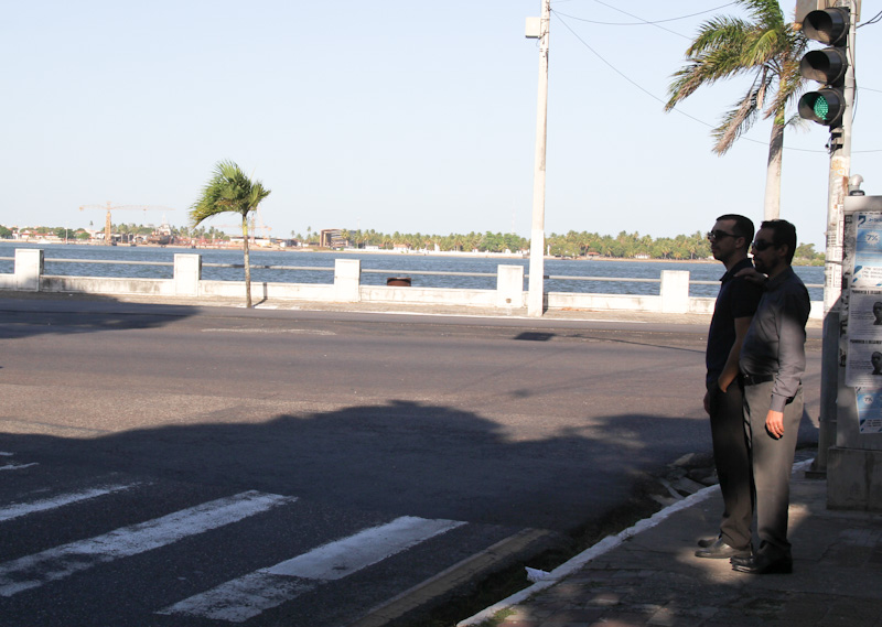 #PraTodoMundoVer Foto de uma avenida, onde há uma faixa de pedestres pintada. Dois homens estão parados na calçada do lado direito