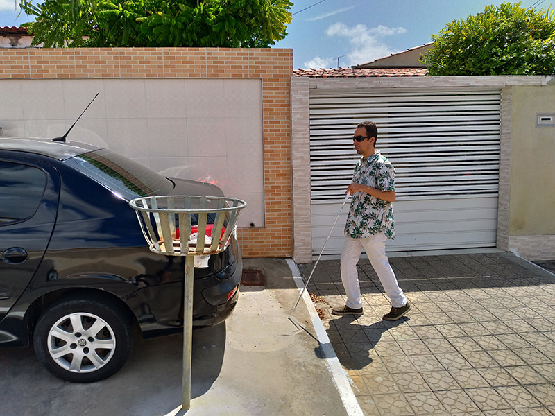 #PraTodoMundoVer Um homem caminhando na calçada em frente a um muro e um portão. Ele segura um begala. À frente dele, há um carro em cima da caçada e uma cesta para lixo.