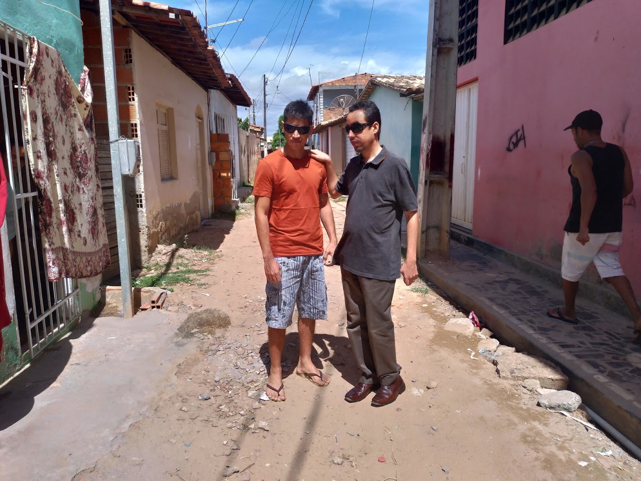 #PraTodoMundoVer Dois homens em pé em uma rua de piçarra. Ao lado há muros de casas.