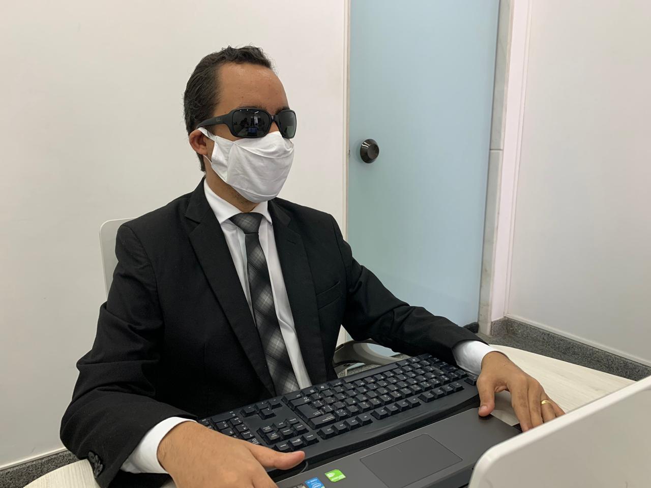 #PraTodoMundoVer Fotografia de Lucas que está sentado usando terno preto, óculos escuros e máscara de proteção facial, com as mãos sobre um teclado de computador. 